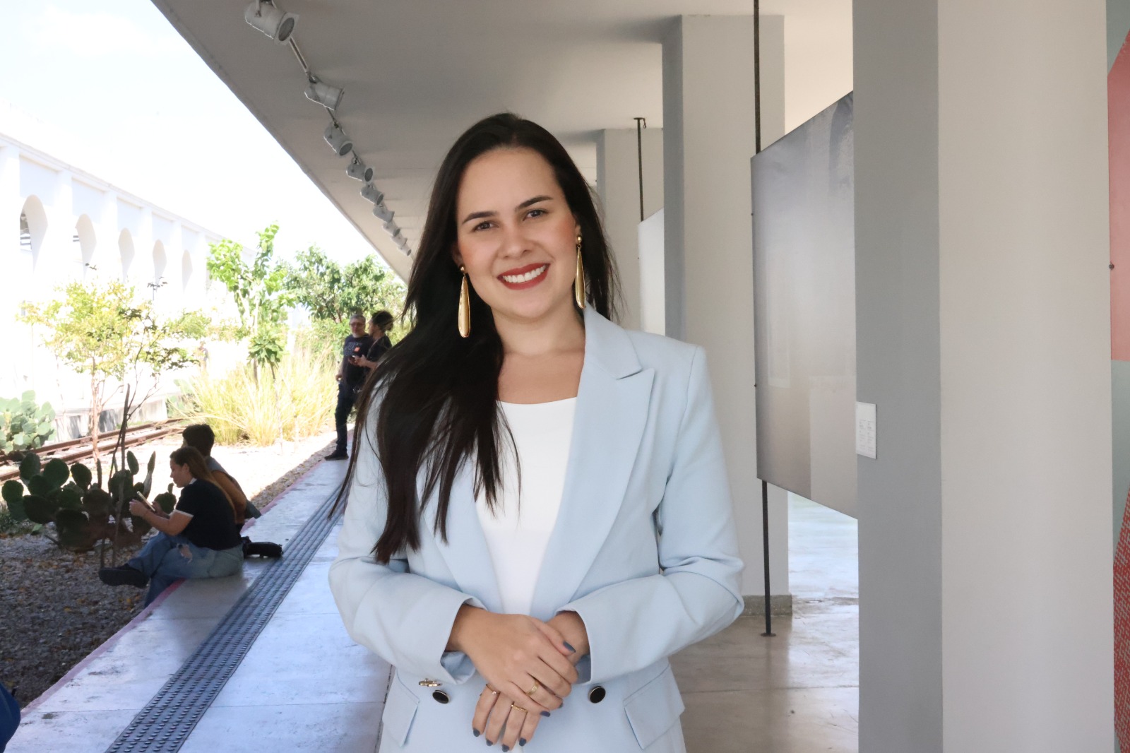 Sabrina Medino posa para a foto; ela é uma mulher branca cis, tem cabelos pretos e veste um blazer azul claro